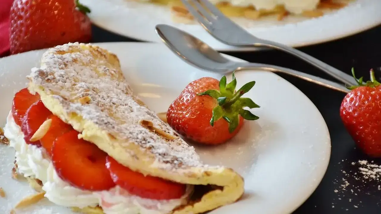 Słodki omlet z bitą śmietaną i truskawkami, posypany cukrem pudrem i płatkami migdałów. Idealny deser na słodko.