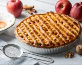 Złocista tarta z jabłkami, udekorowana kratką z ciasta, obok czerwonych jabłek i cynamonu.