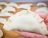 Dłoń trzyma surowe pierogi z serem, gotowe do gotowania. Na drewnianej desce widać więcej pierogów.