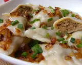 Pyszne pierogi z mięsem, okraszone skwarkami i szczypiorkiem, w białej misce.