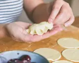 Dłonie formują ciasto na pierogi, tworząc pyszne danie. Na blacie leżą okrągłe placki z ciasta, a obok miseczka z wiśniami.