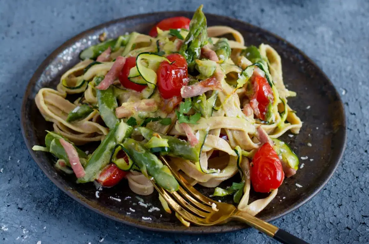 Pyszna carbonara ze szparagami, pomidorkami i cukinią na talerzu. Złoty widelec czeka, by spróbować tego wiosennego dania.