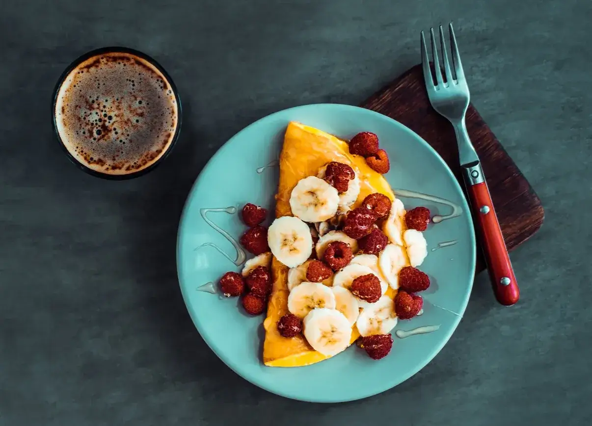 Słodki omlet z bananami i malinami, polany miodem, obok filiżanka kawy. Idealne śniadanie.