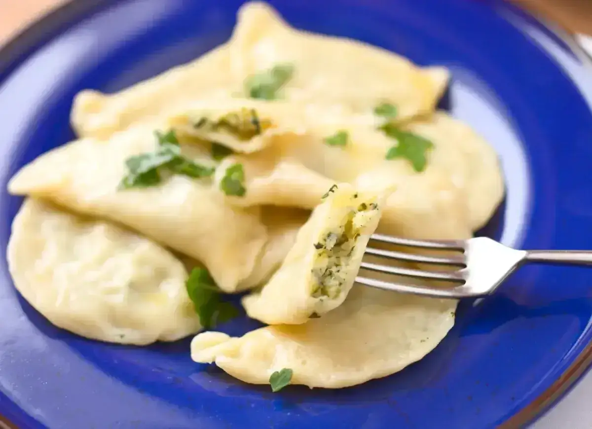 Pyszne pierogi ze szpinakiem, polane masłem i posypane natką pietruszki, na talerzu. Jedna sztuka jest nadziana zielonym farszem.