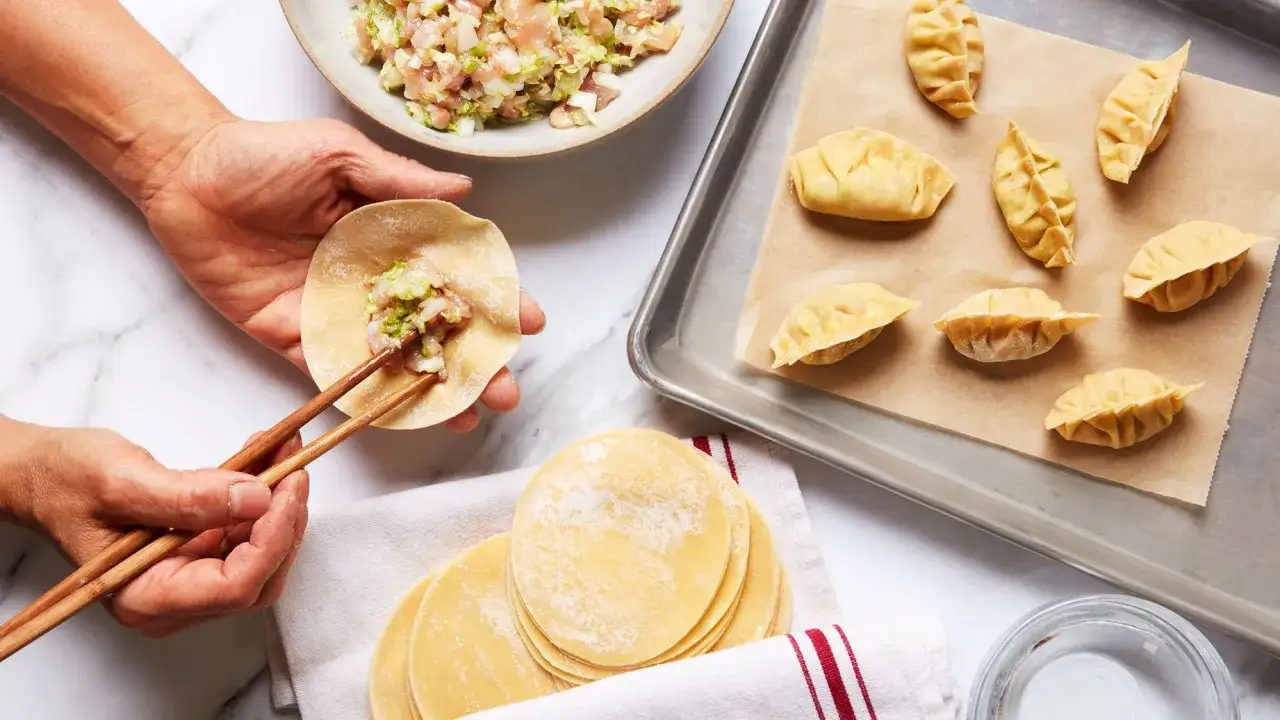 Ręce formują pierożki dim sum, nakładając farsz na ciasto pałeczkami. Obok gotowe pierożki na blasze.